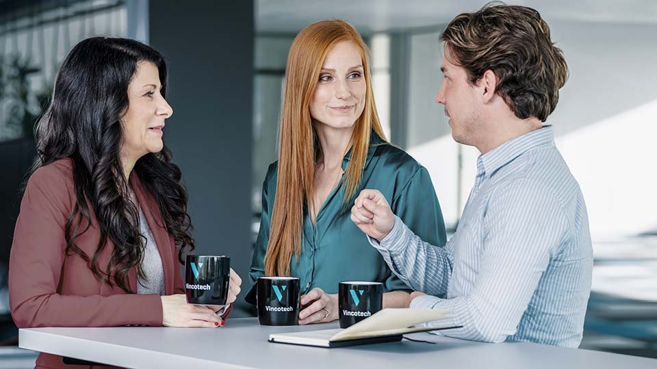 Three people having a conversation at a table with cups and a notebook, indoors with modern decor.