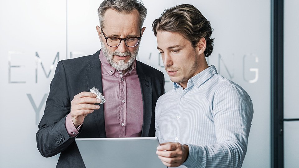 Two men reviewing information on a tablet; one holds a power module, wearing business attire in a modern office.