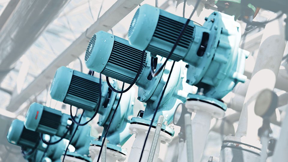 Row of industrial blue electric motors with cooling fins, mounted on white pipes in a factory setting.