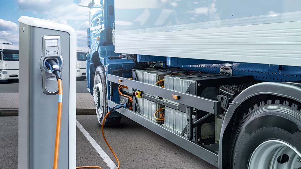 Electric truck charging at station, with visible battery packs and connected orange cable. Background shows more trucks and a cloudy sky.