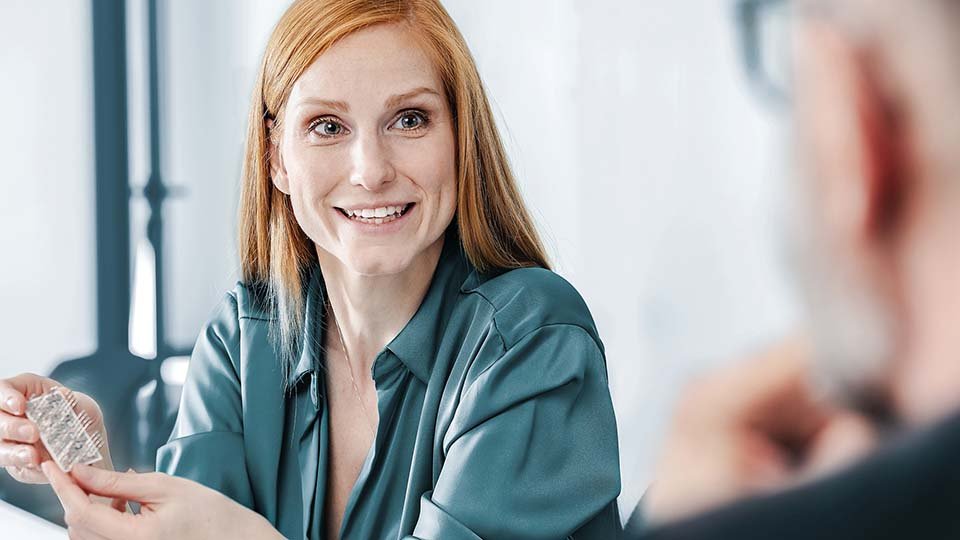 Smiling woman with red hair holds up a power module, sitting at a table, in conversation with a blurred person in the foreground.