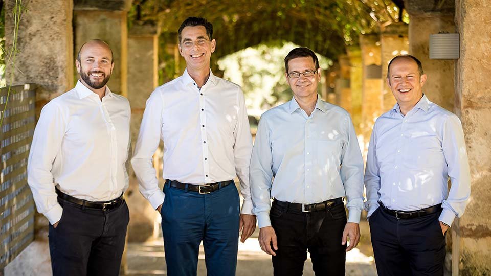 Four men in white shirts standing and smiling, under a stone archway with greenery above, in a sunlit outdoor setting.
