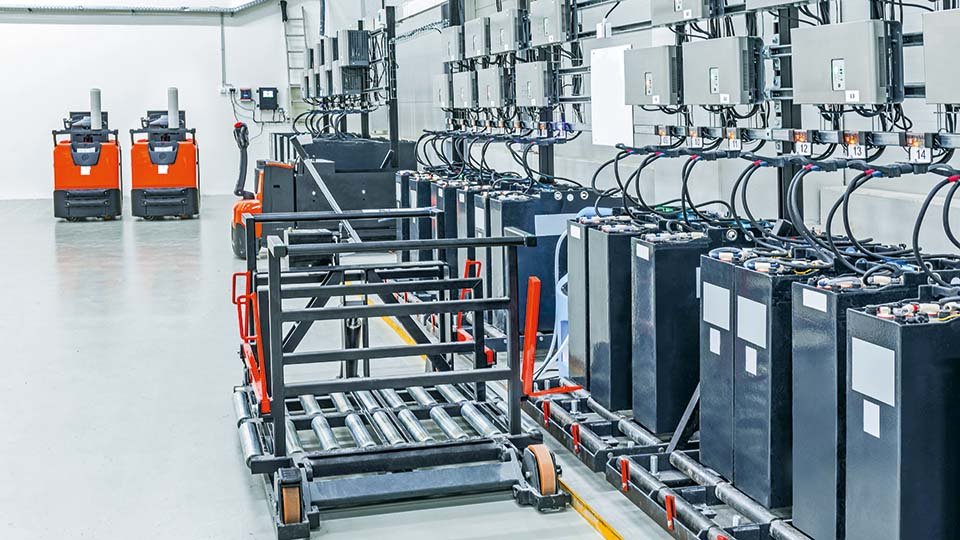 Industrial battery charging room with multiple charging units, black batteries, and orange forklifts against a clean white wall.