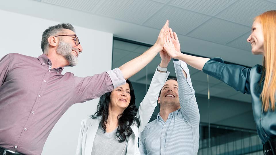 Four colleagues smiling and giving a group high-five in an office setting.