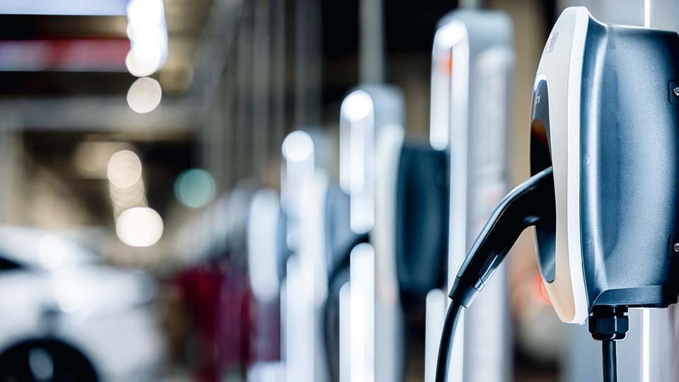 Close-up of an electric car charging station with a plugged-in cable, set against a blurred industrial background.