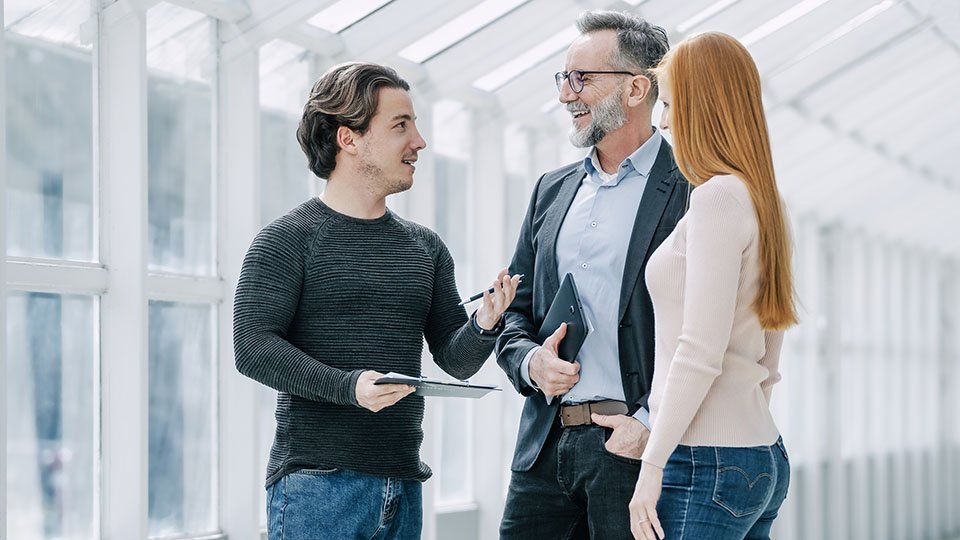 Three people discussing in a bright, glass-walled hallway. One holds a tablet, while the others engage with smiles and attentive gestures.