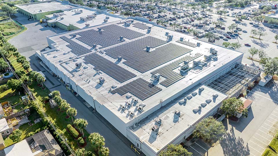 Aerial view of a large commercial building with solar panels on the roof, surrounded by parking lots and greenery.