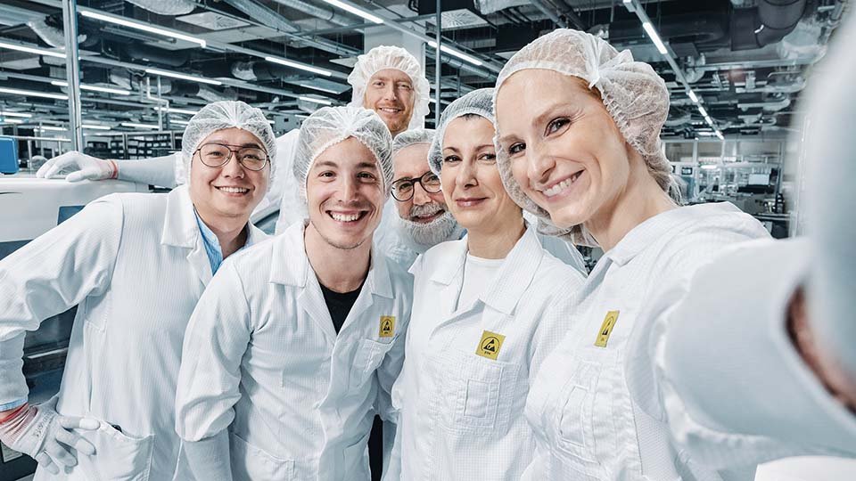 A group of six people wearing white lab coats and hairnets smile for a selfie in an industrial lab setting.