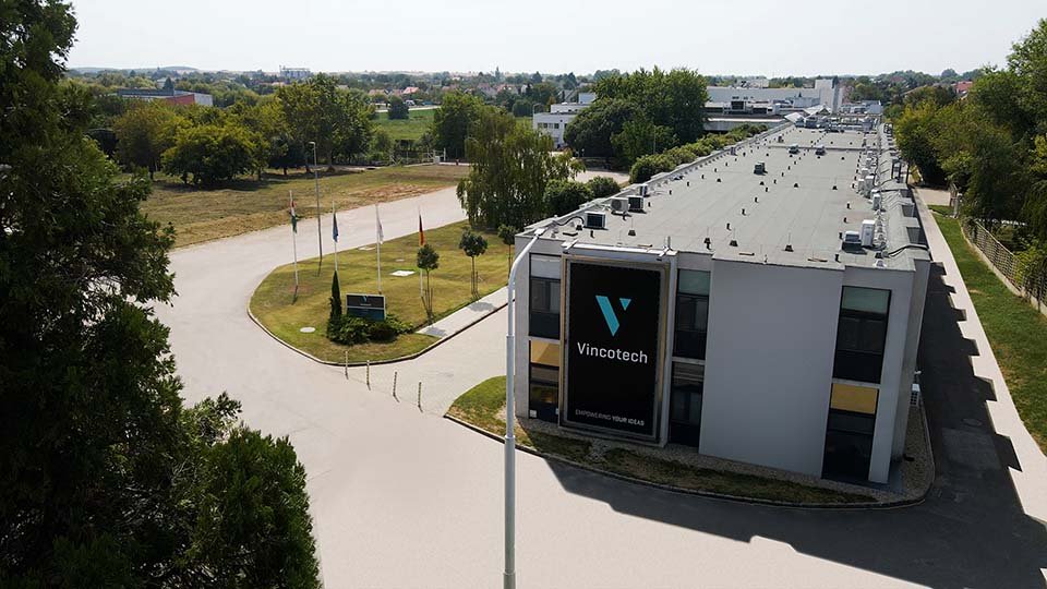 Aerial view of the Vincotech building with a sign reading "Vincotech." Surrounding trees and flags line a curved path.