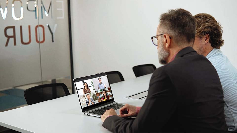 Two men sit at a table, participating in a video call on a laptop, with a glass wall showing partial text in the background.