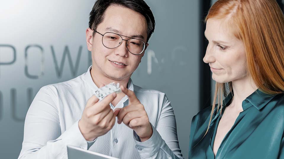 Two people discuss a small mechanical part, one pointing to it while the other listens attentively in a modern office setting.