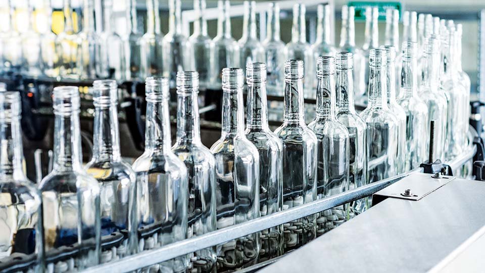 Rows of clear glass bottles on a conveyor belt in a factory setting, ready for filling or packaging.