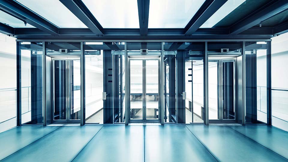 Modern glass elevators with a sleek design, featuring transparent doors and clean lines, situated in a bright, spacious building interior.