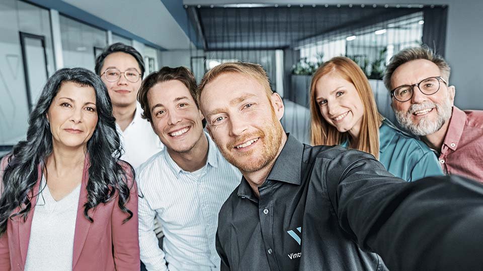 A diverse group of six people smiling for a selfie in a modern office setting.