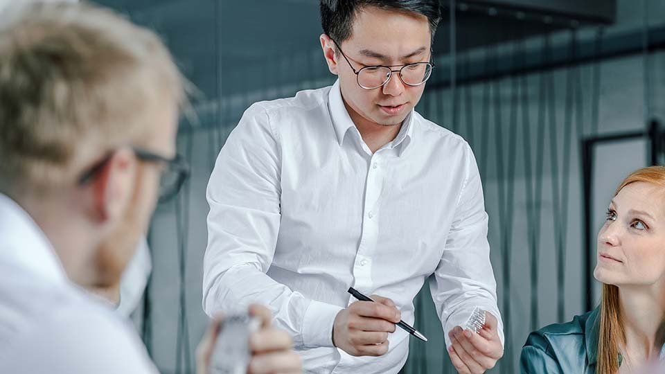 A man in glasses, holding a pen and a power module, explains to two colleagues in an office setting.
