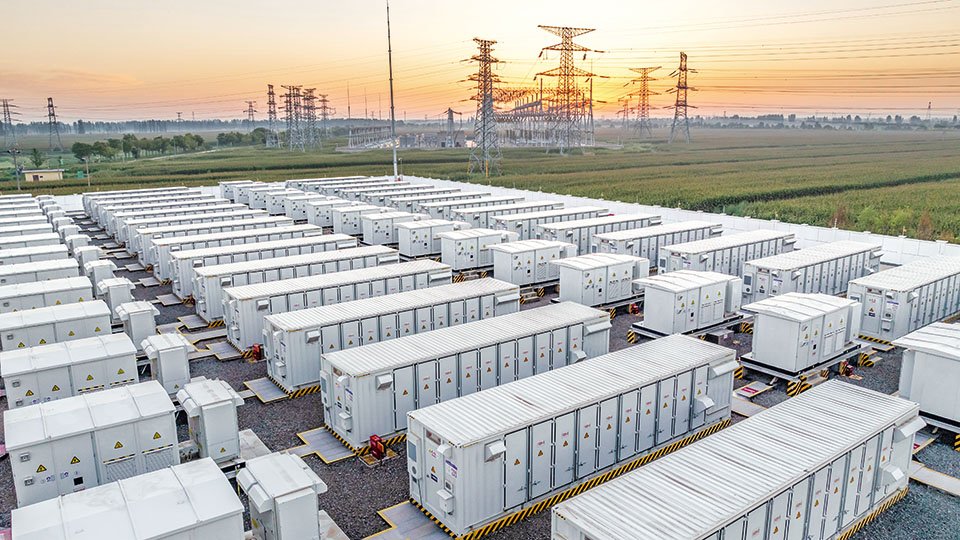 Rows of white energy storage containers in a field with electrical towers, under a sunrise sky.