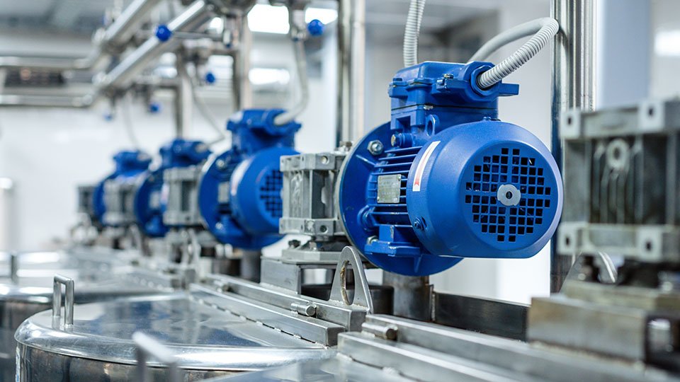 Row of blue industrial electric motors mounted on metal frames, inside a manufacturing plant with pipes above.