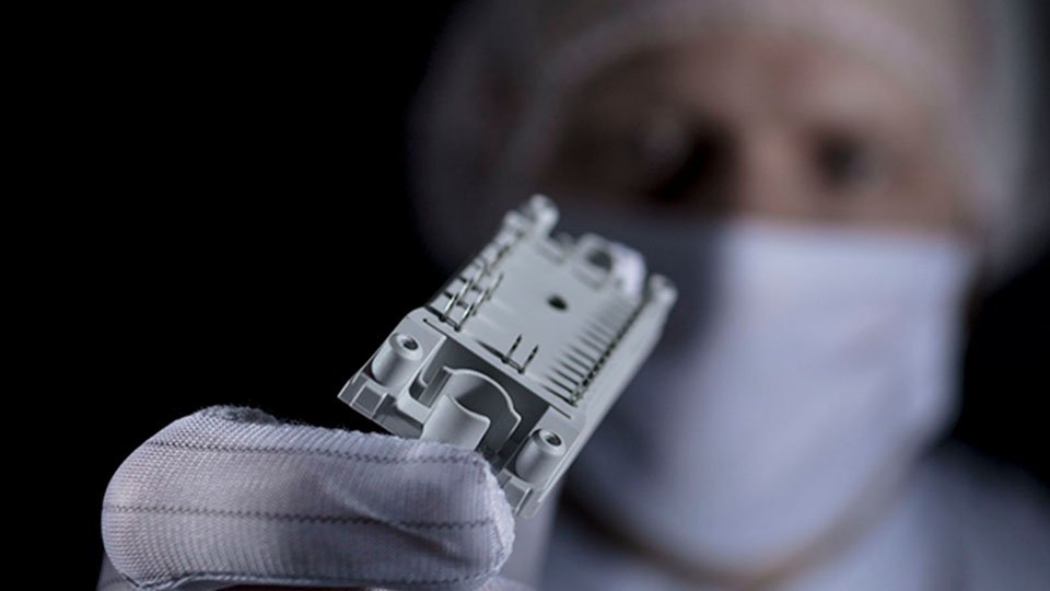 A person wearing gloves and a mask holds a power module in a precise manner against a dark background.