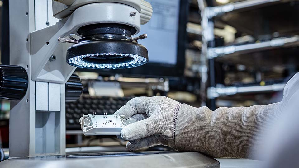 Gloved hand holding a power module under a microscope with LED light in an industrial setting.