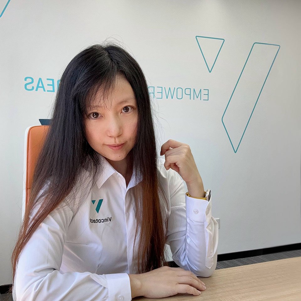 Anna sits at a desk, wearing a white shirt with a logo, in a modern office setting with geometric wall art.