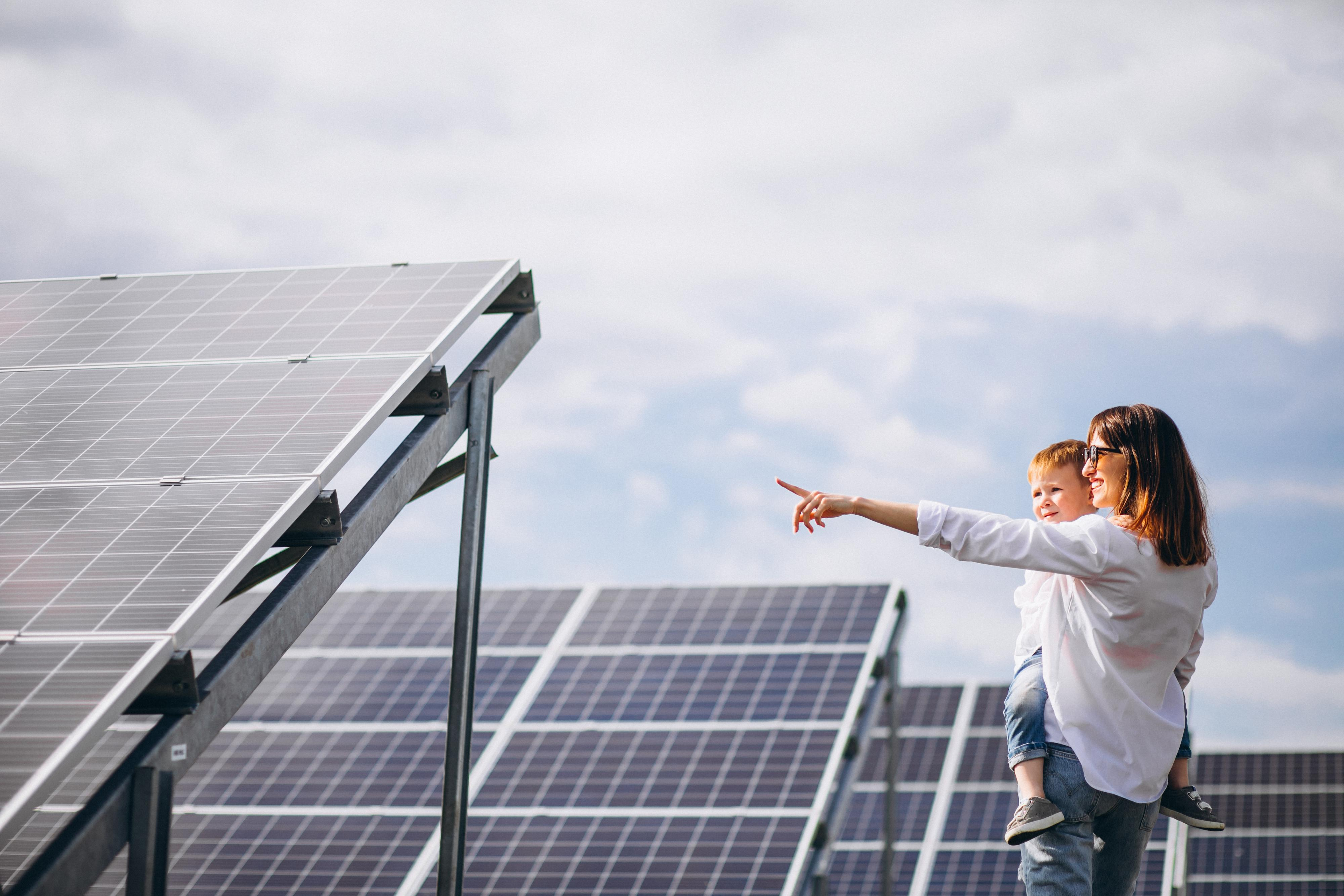 A woman in sunglasses holds a child, both pointing left at an array of solar panels under a partly cloudy sky.