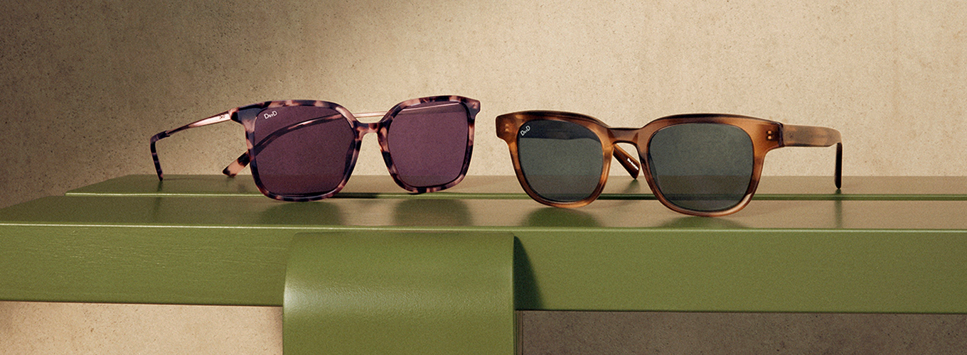 Two pairs of sunglasses on a green surface: pink tortoiseshell with purple lenses, and brown tortoiseshell with dark grey lenses.
