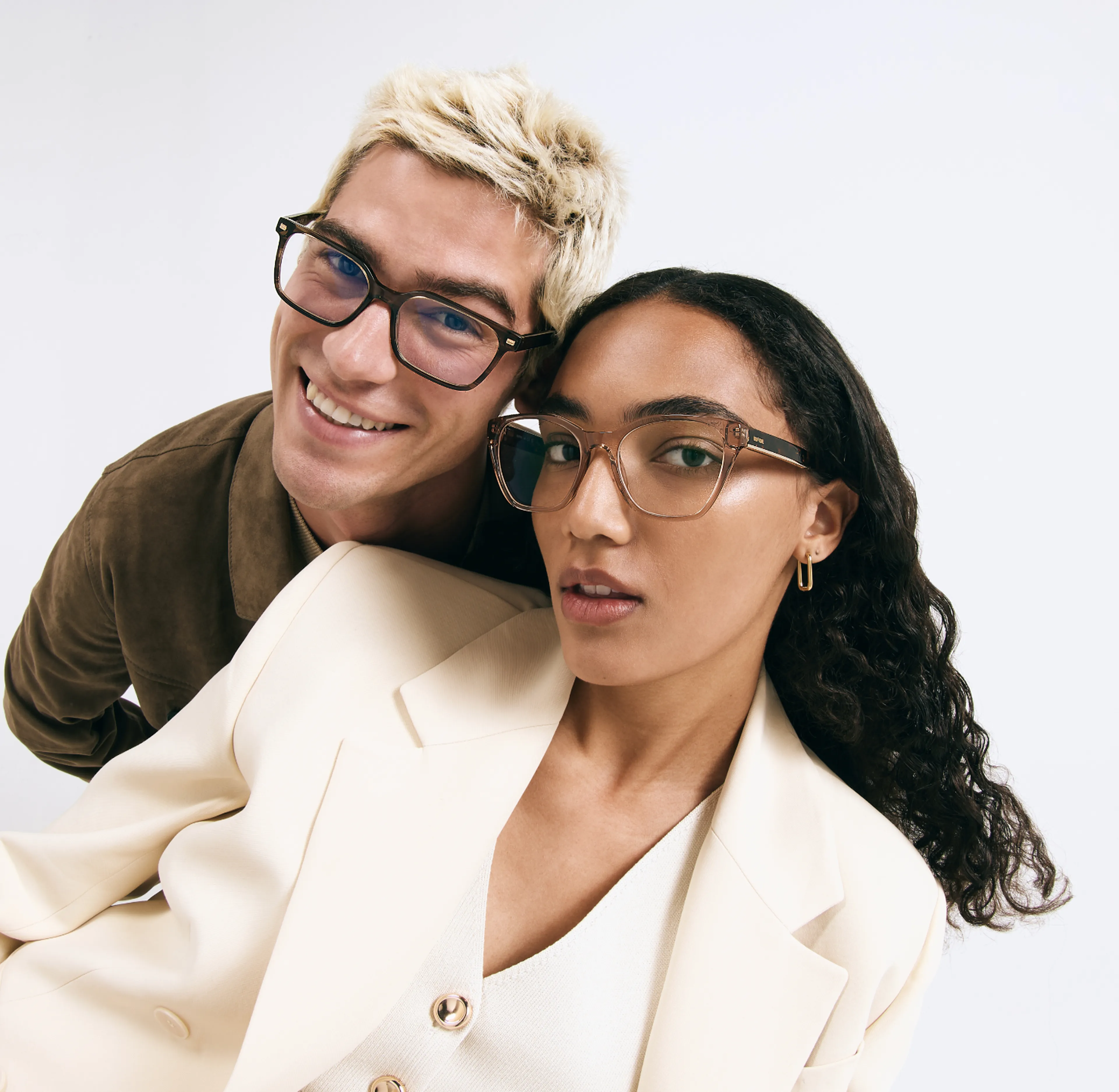 A smiling man with blonde hair and a woman with dark curly hair, both wearing stylish eyeglasses, pose against a white background.