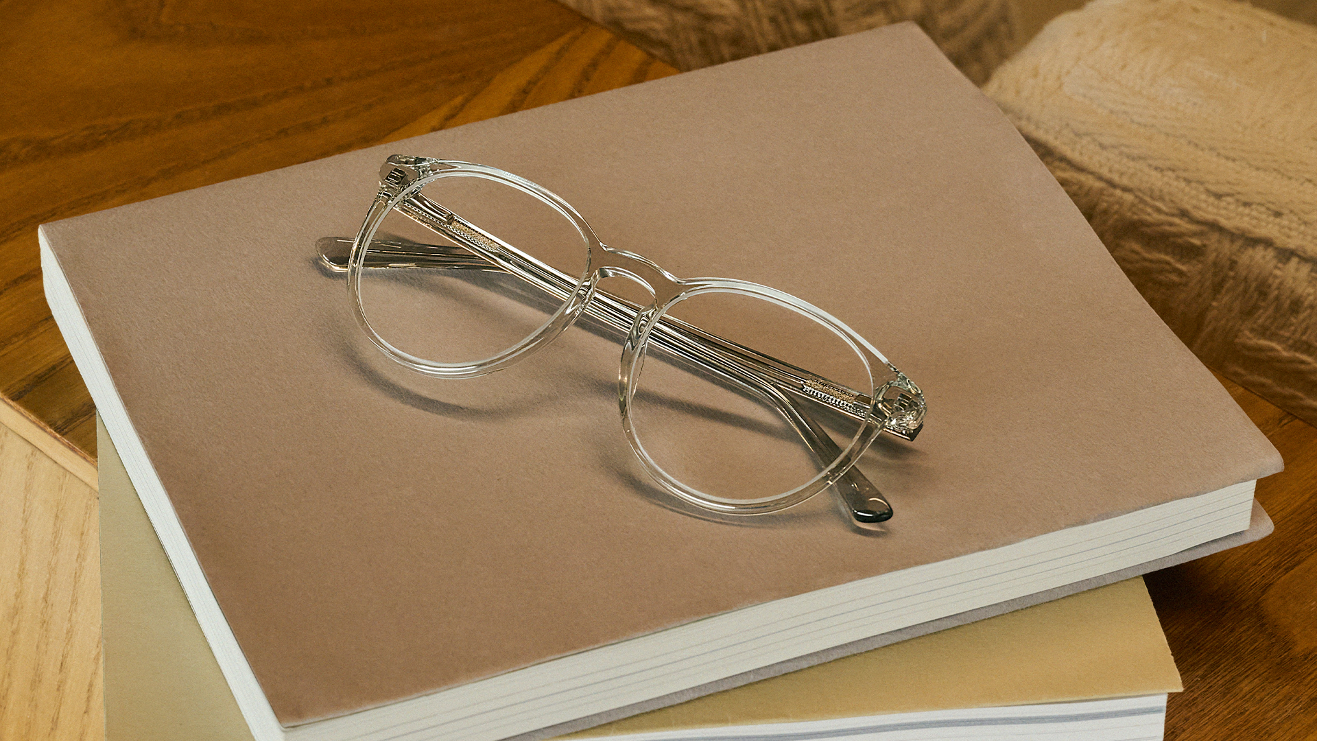 Clear-framed glasses rest on a brown notebook, part of a stack, on a wooden surface with light yarn.