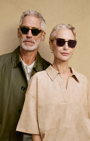 Older, grey-haired couple in sunglasses. Man in green coat, woman in beige top, against a beige wall.