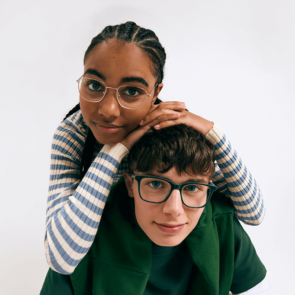 Two smiling teens in glasses. A girl with braids and gold frames hugs a boy with green frames from behind, looking at the camera.