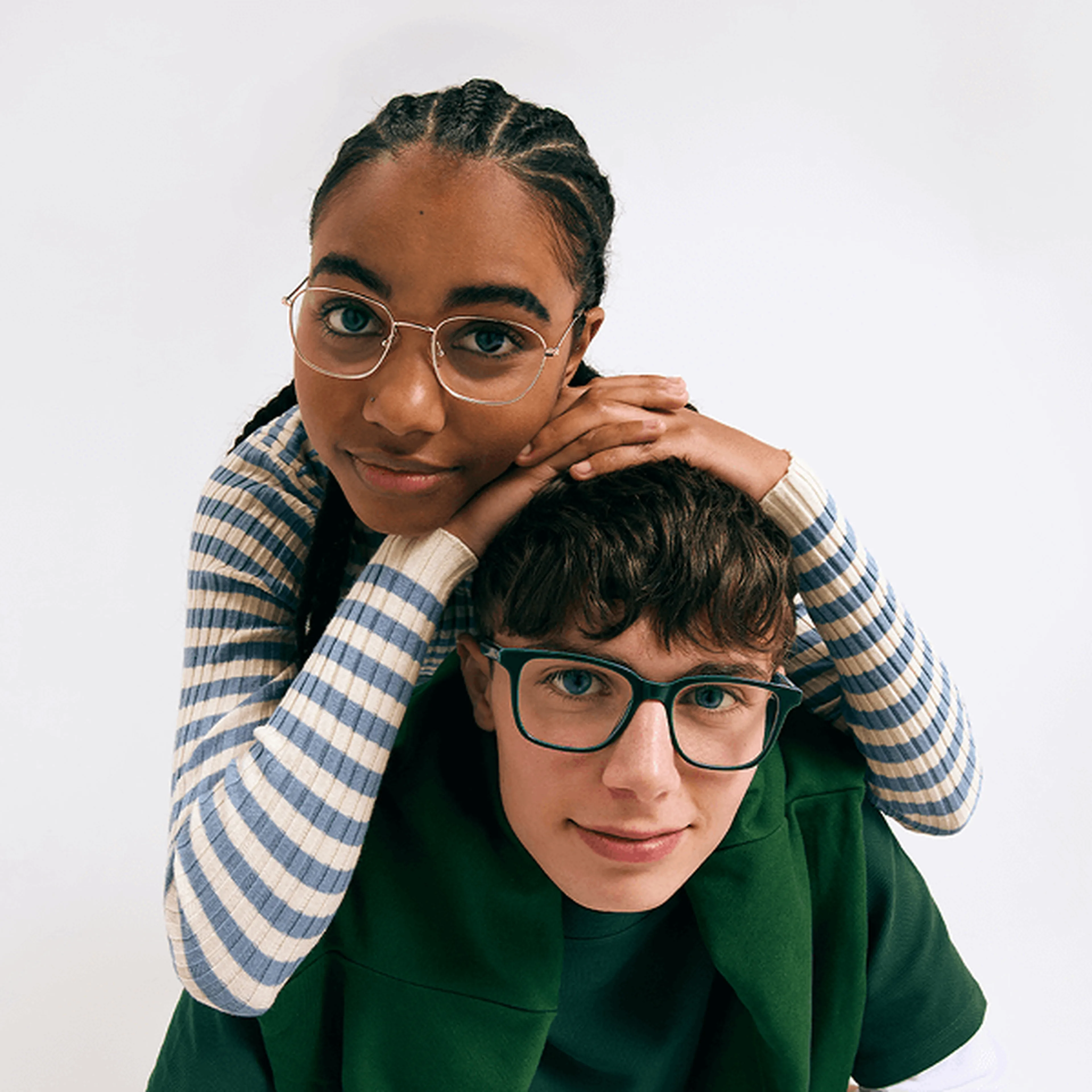 Two smiling teens in glasses. A girl with braids and gold frames hugs a boy with green frames from behind, looking at the camera.