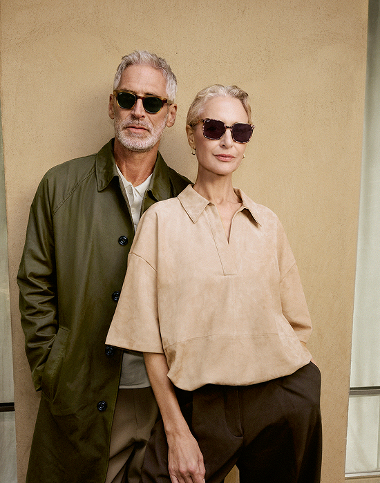 Older, grey-haired couple in sunglasses. Man in green coat, woman in beige top, against a beige wall.