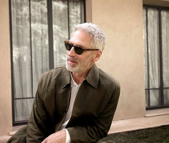 A man with gray hair and beard, wearing tortoise-shell sunglasses and a green jacket, looks right. Building with windows in background.