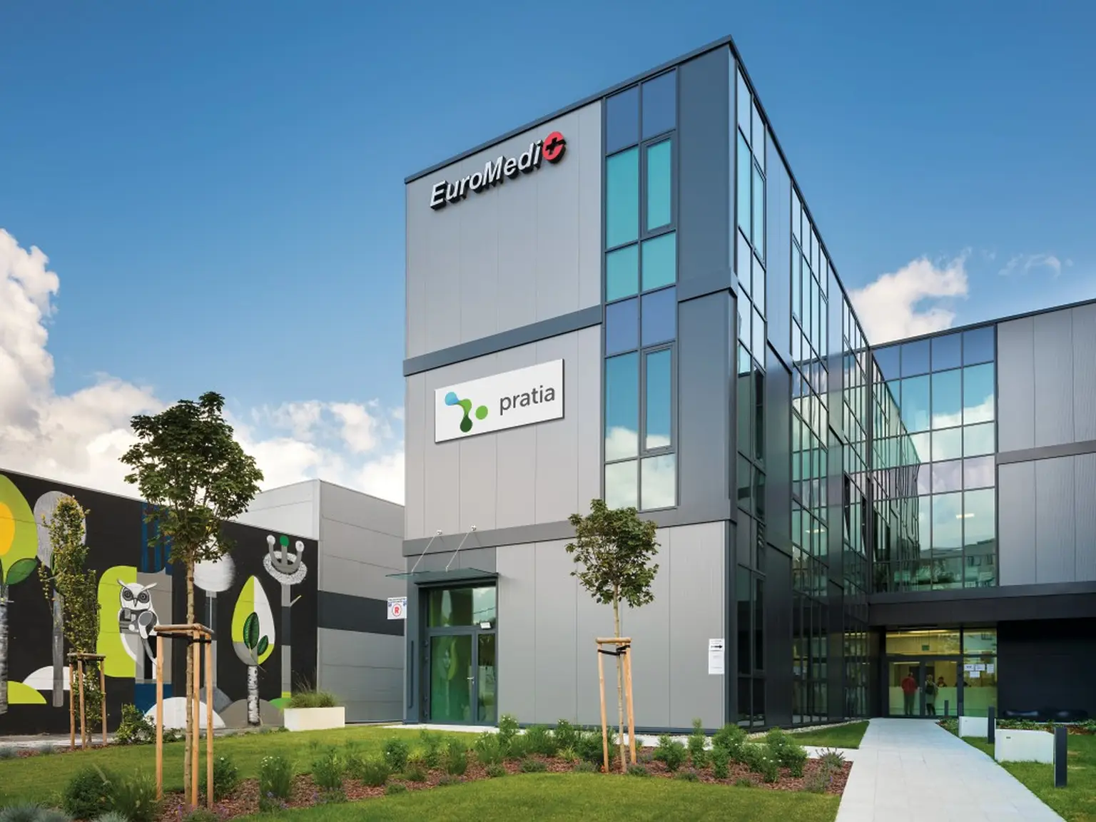 Modern building with glass facade, labeled 'pratia' and 'EuroMedic.' Features greenery and a mural of stylized trees and an owl.