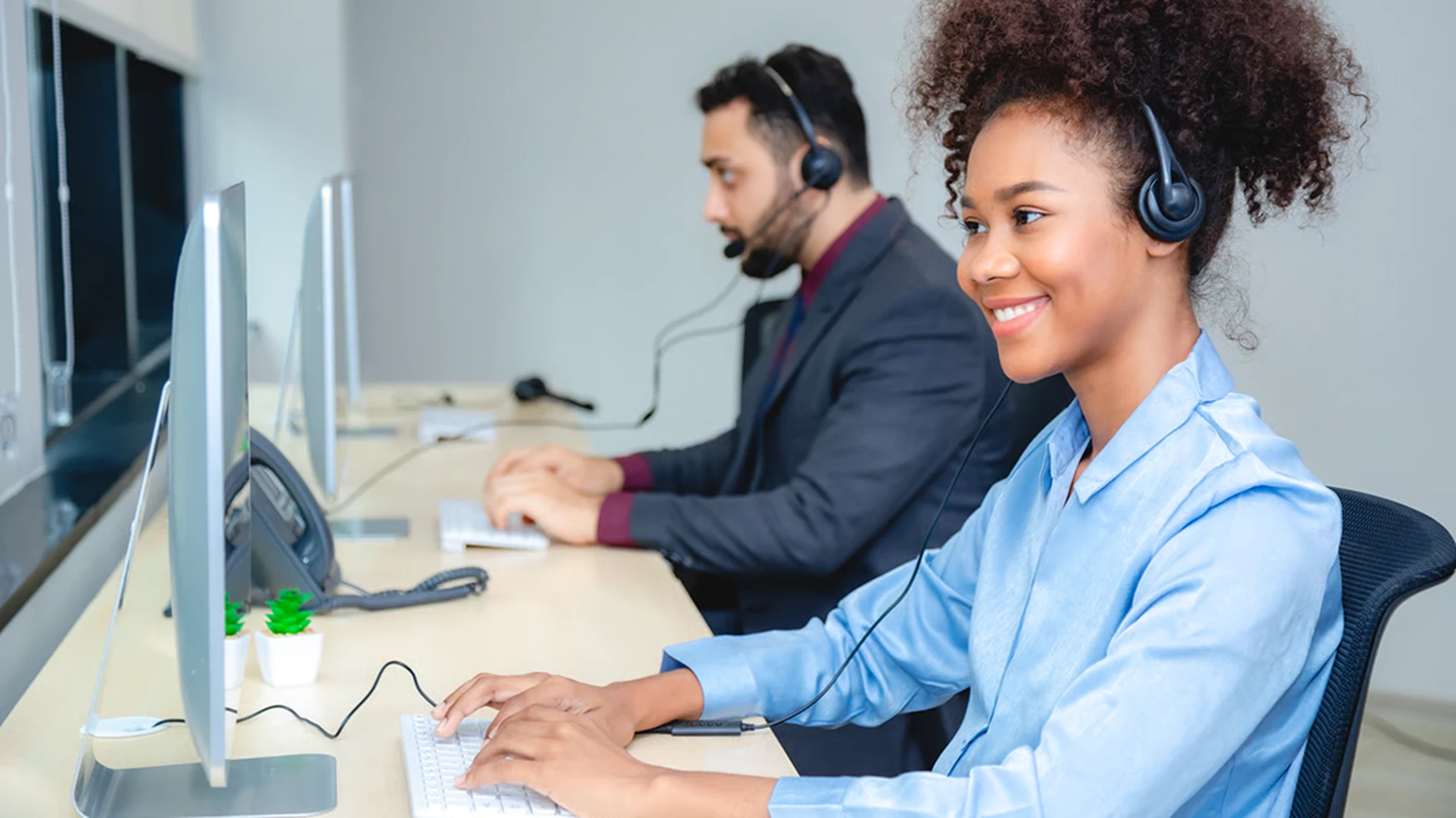 Two customer service representatives wearing headsets work at computers in a bright office. A small plant decorates the desk.