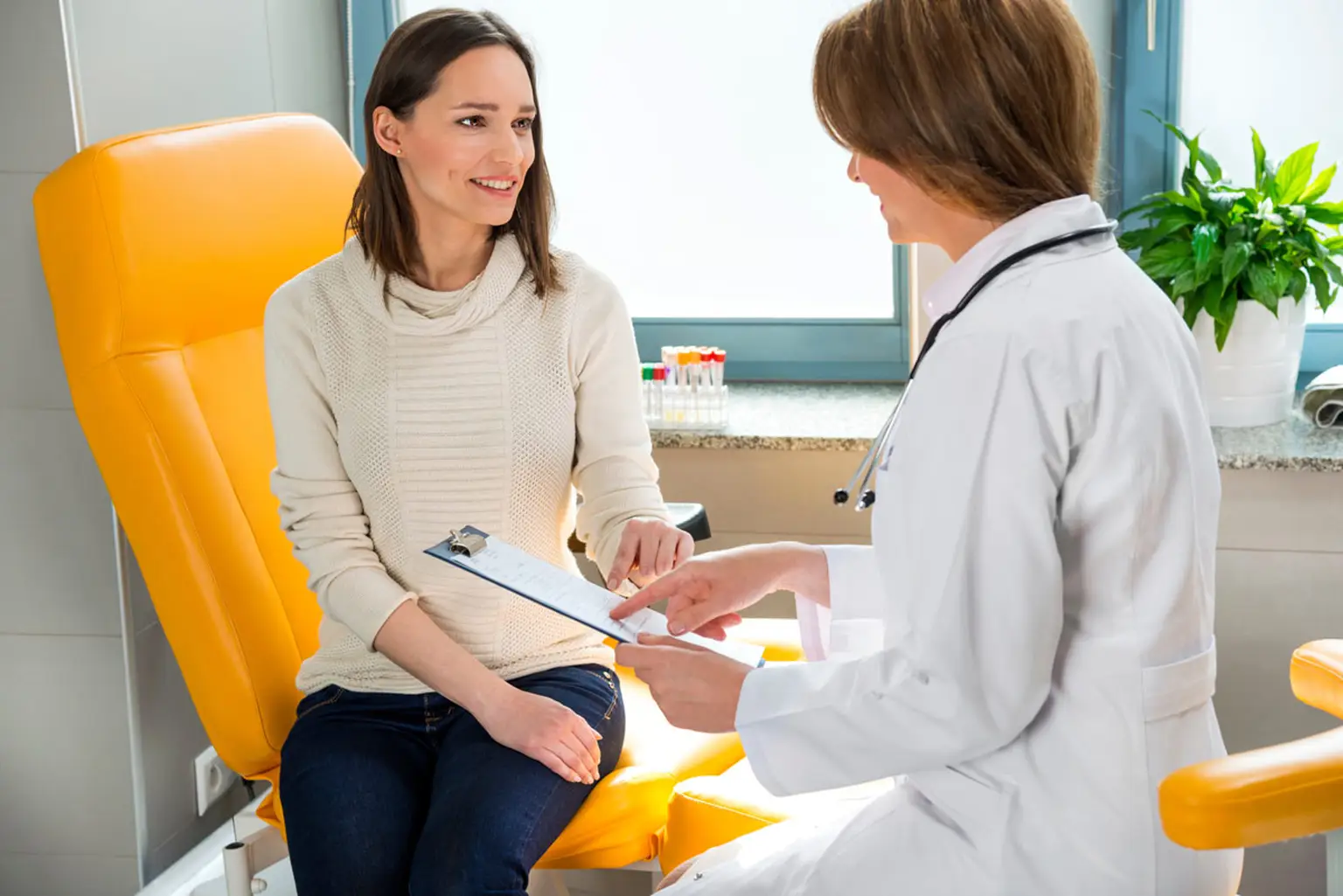 A doctor and patient at a Pratia research site discuss medical details in a bright office. The patient is seated on a yellow chair, holding a clipboard.
