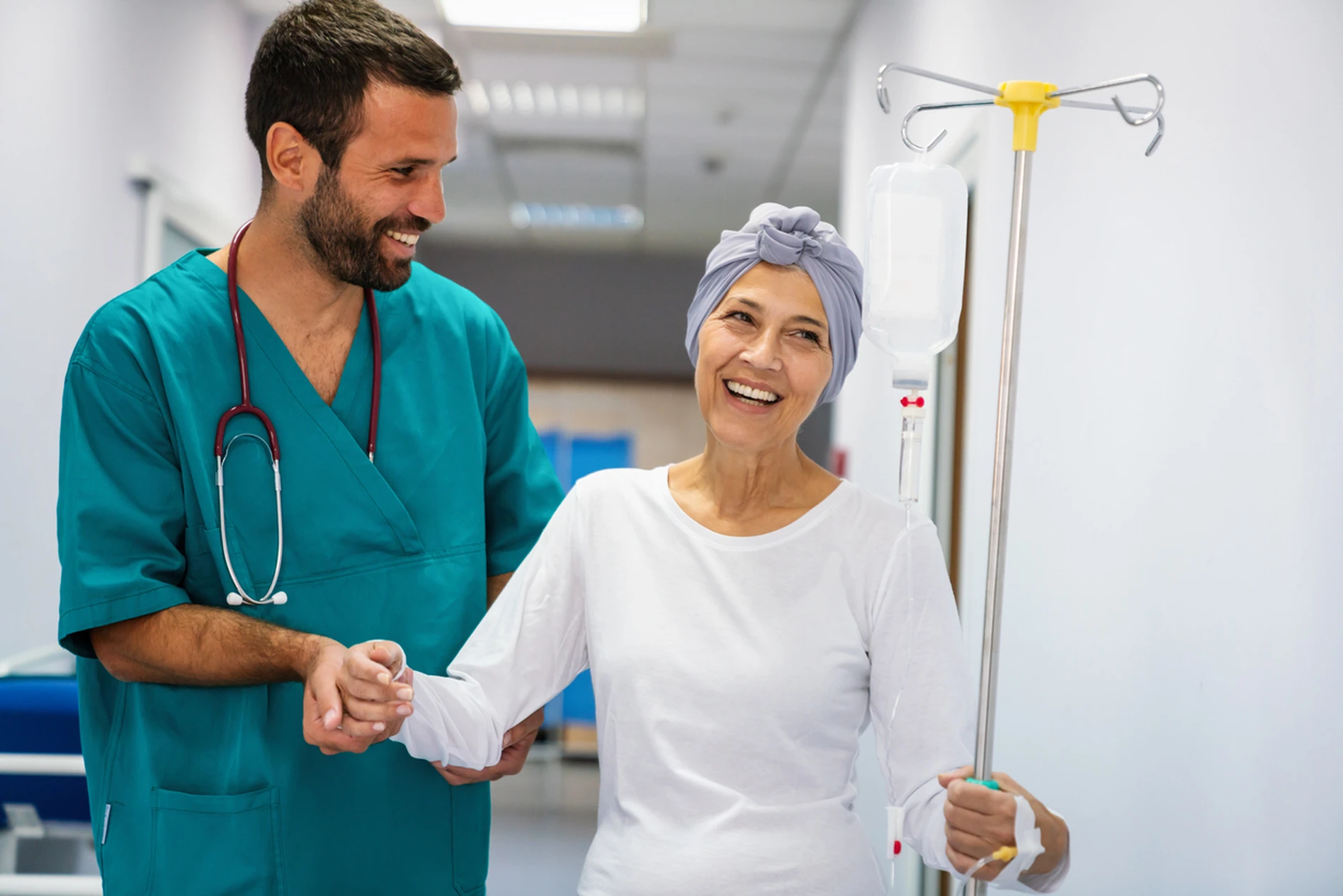 Healthcare professional assists smiling patient with headscarf holding an IV pole in a hospital hallway.