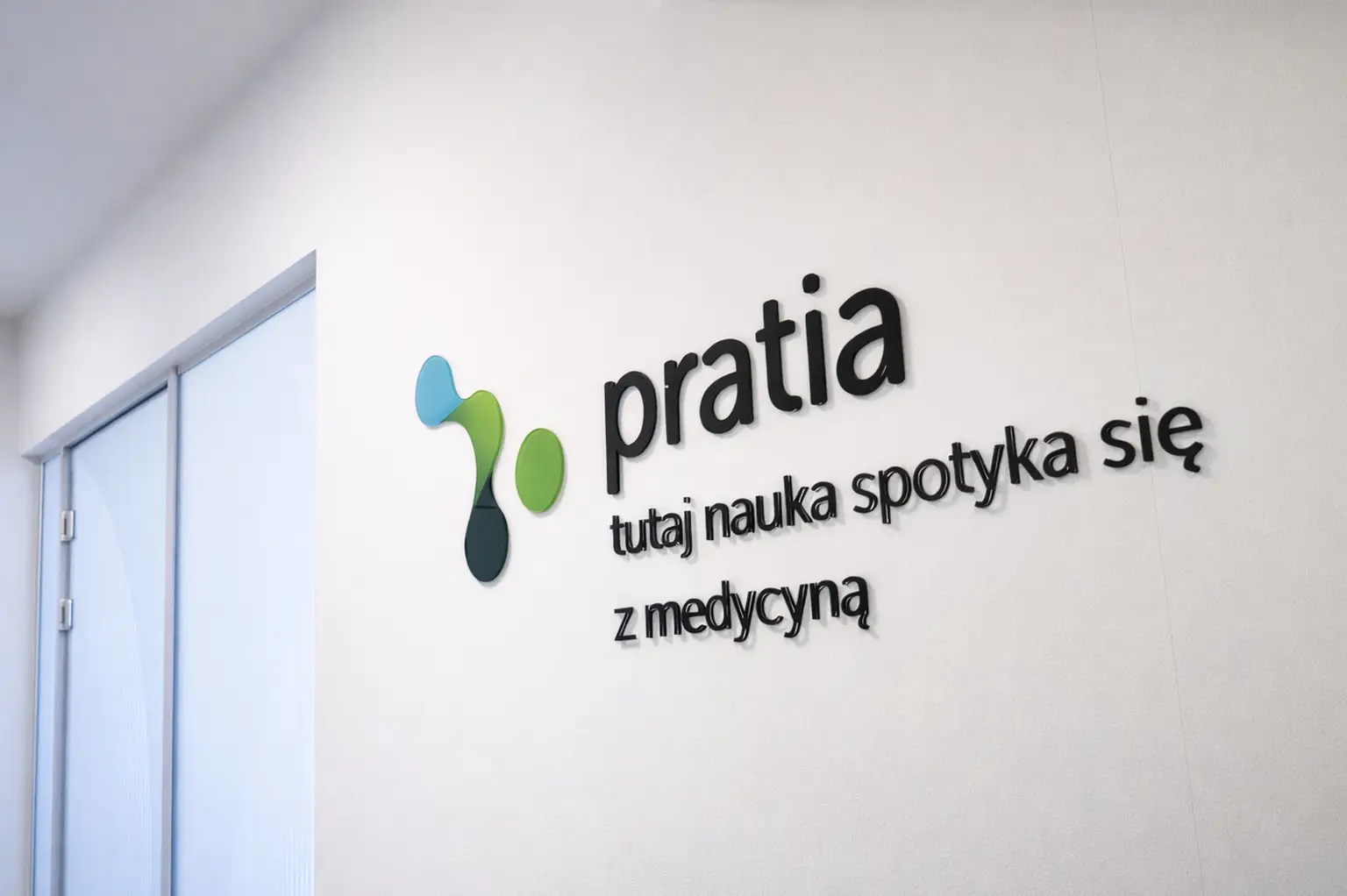 Wall with "pratia" and "tutaj nauka spotyka się z medycyną" text beside a blue, green, and dark green abstract logo.