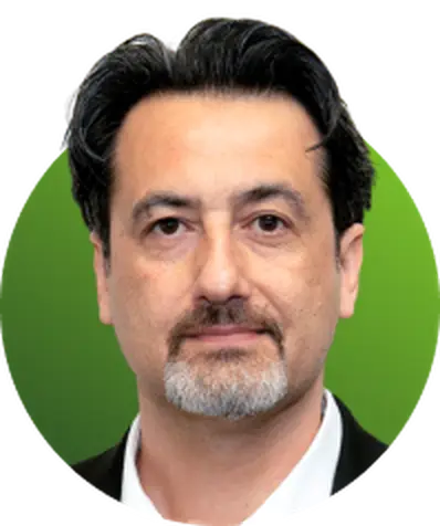 Man with dark hair and a grey beard, wearing a black suit jacket and white shirt, posed against a green gradient background.