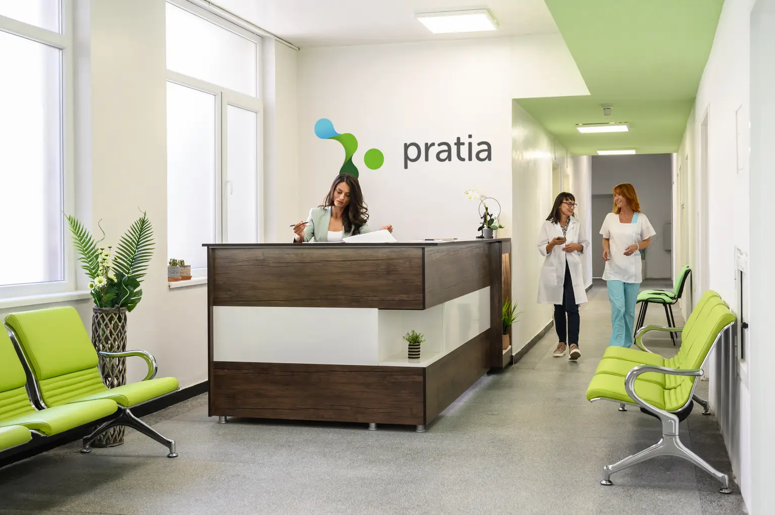 Modern medical office lobby at one of Pratia's Research Sites with a receptionist at a wooden desk and two professionals walking in the hallway, bright green seating.