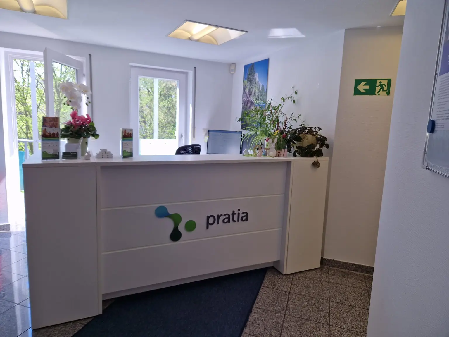 Reception desk at a Pratia research site  with flowers and brochures, labeled "pratia," in a well-lit office space with windows and emergency exit sign.