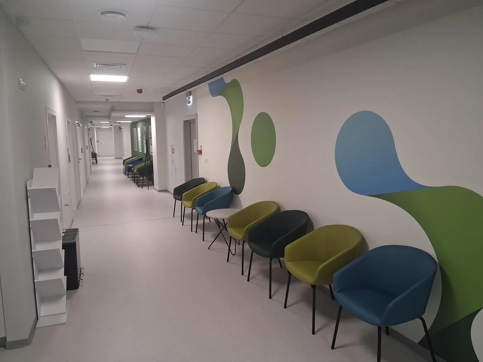 Hospital corridor at a Pratia research site with colorful chairs and abstract wall art on the right, leading to doors and windows in the distance.