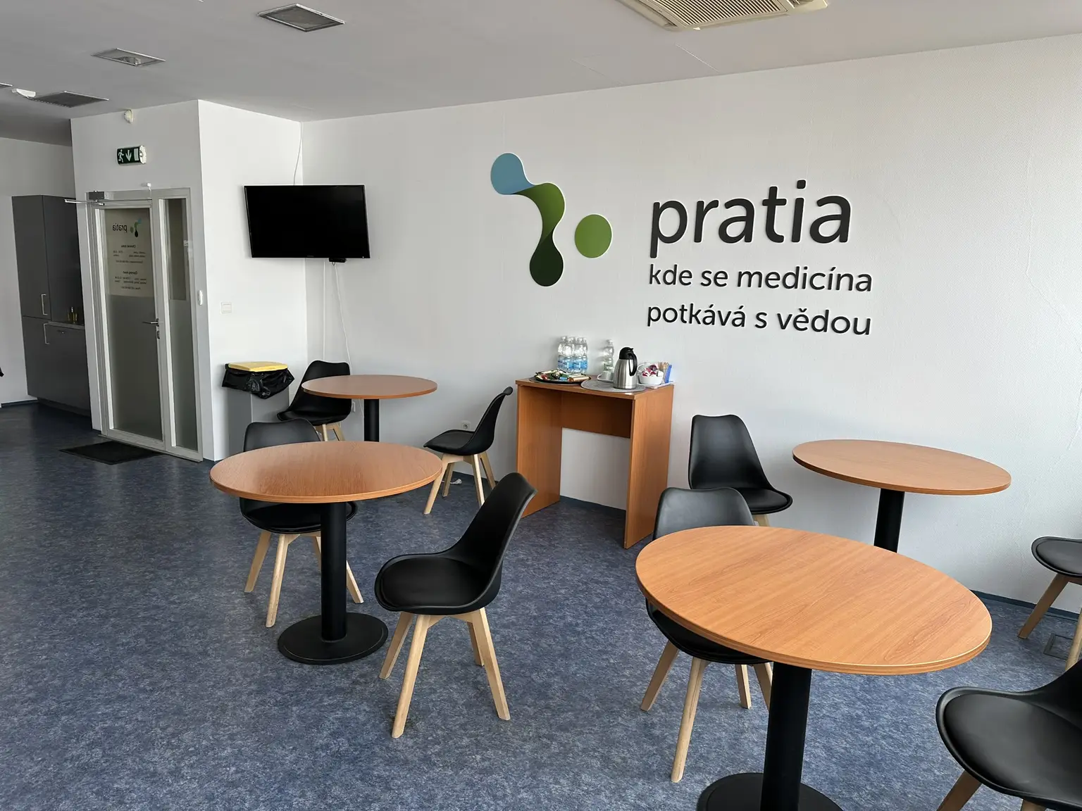 Modern waiting area at a Pratia research site in Czechia with round tables, black chairs, a TV on the wall, and a small refreshment table. "Pratia" logo on the wall with a claim in Czech language