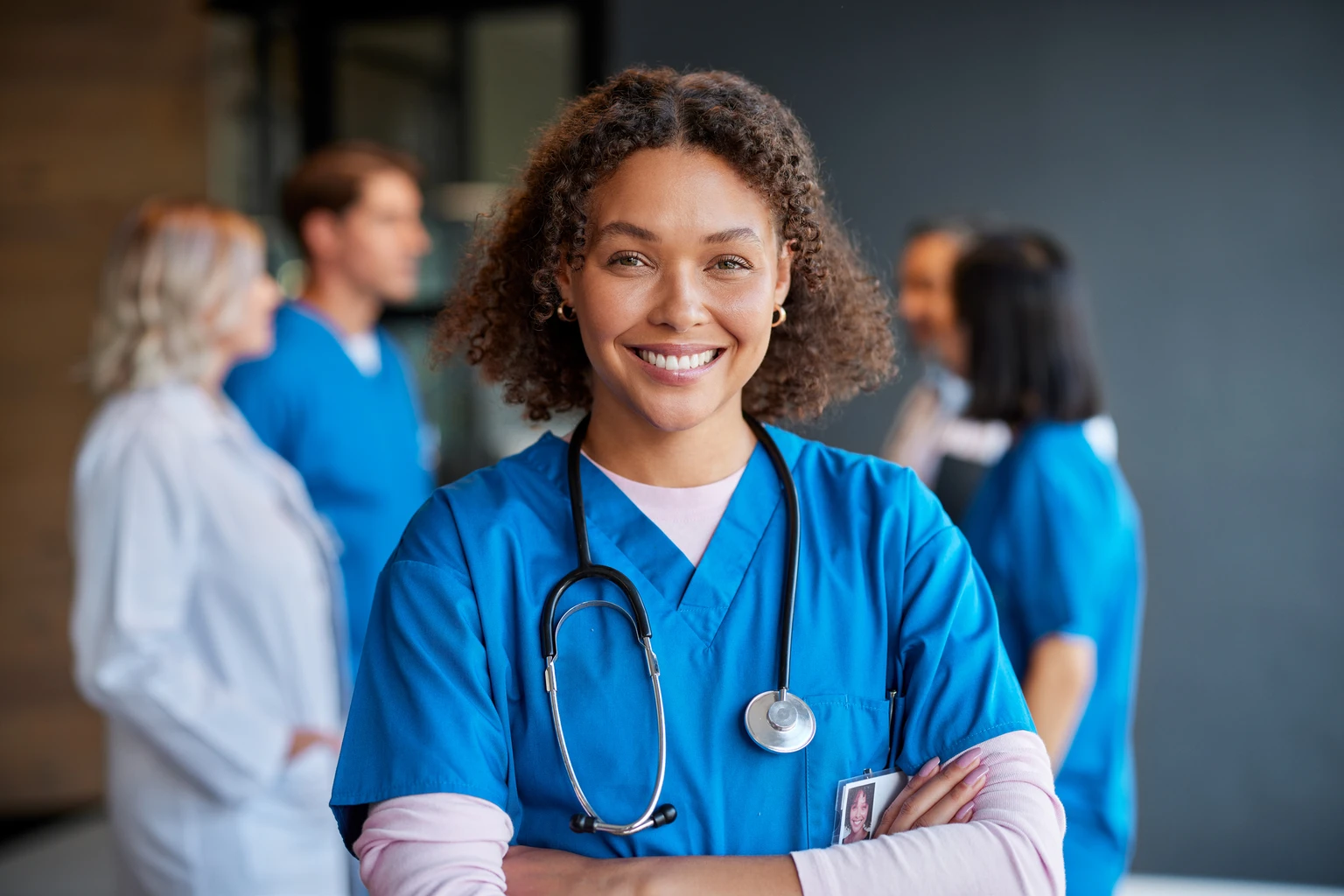 Smiling healthcare professional in blue scrubs with a stethoscope, standing confidently with colleagues in the background.