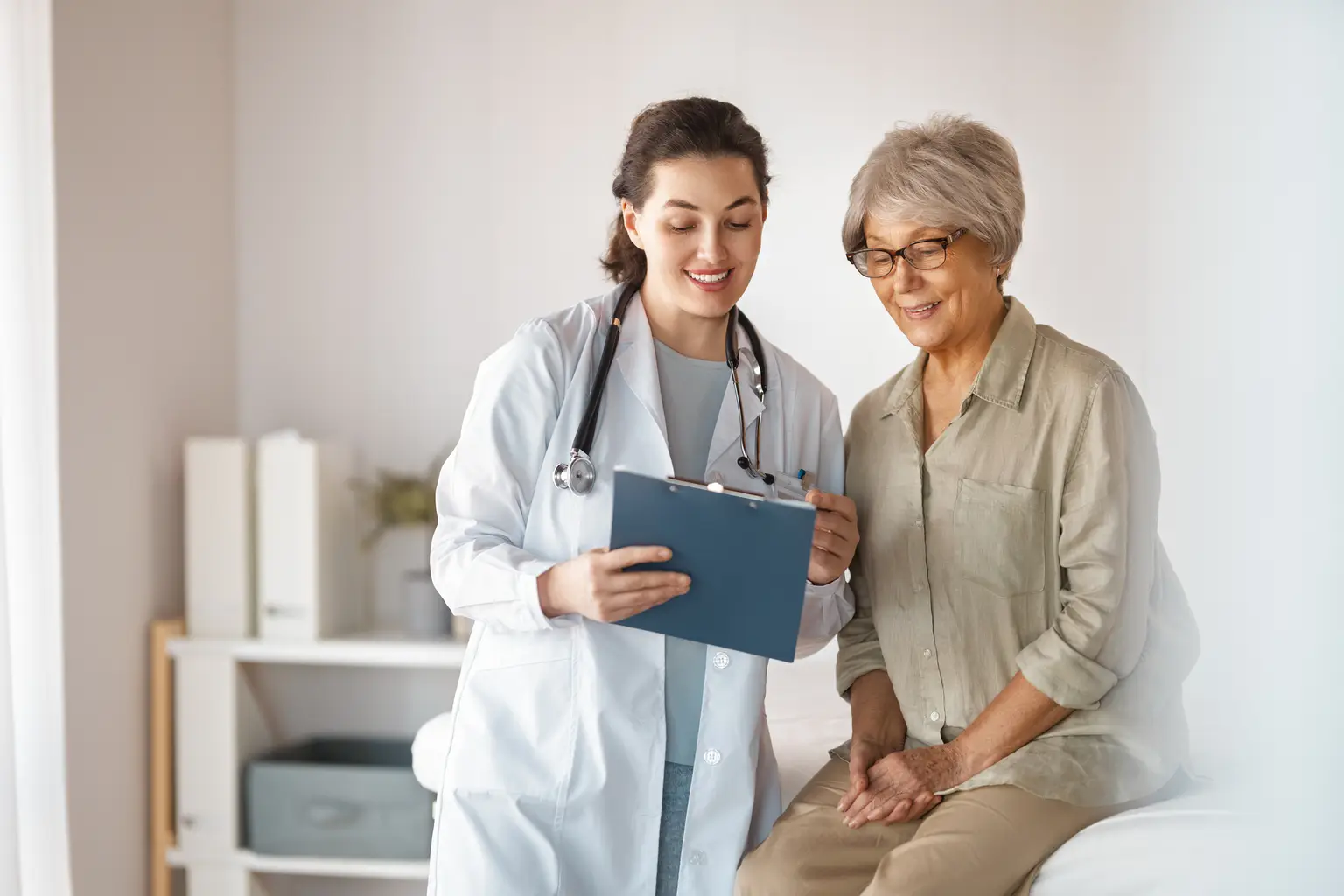 Doctor discussing notes on a clipboard with an elderly patient in an examination room. Both are smiling.
