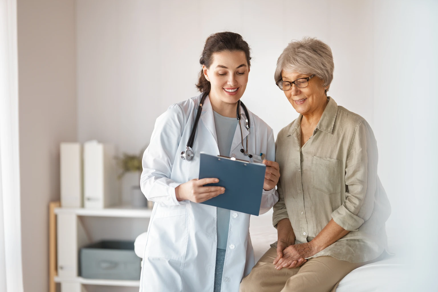 Doctor discussing notes on a clipboard with an elderly patient in an examination room. Both are smiling.