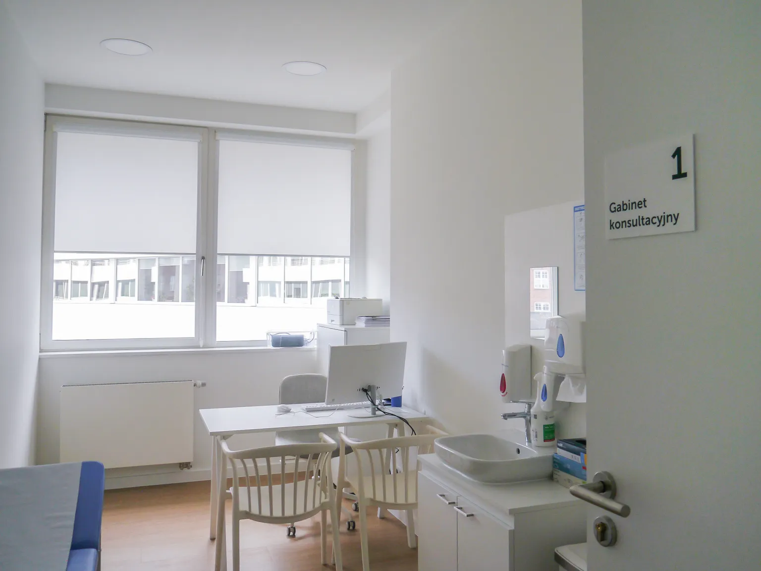 A clean consultation room at Pratia research site with a desk, chairs, computer, and sink. The door has a sign labeled "Gabinet konsultacyjny."