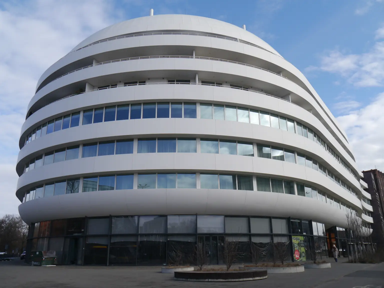 Modern, round, multi-story building - Ovo in Wrocław Poland, houses Pratia Wrocław,  with white, curved facade and numerous windows under a blue sky with clouds.