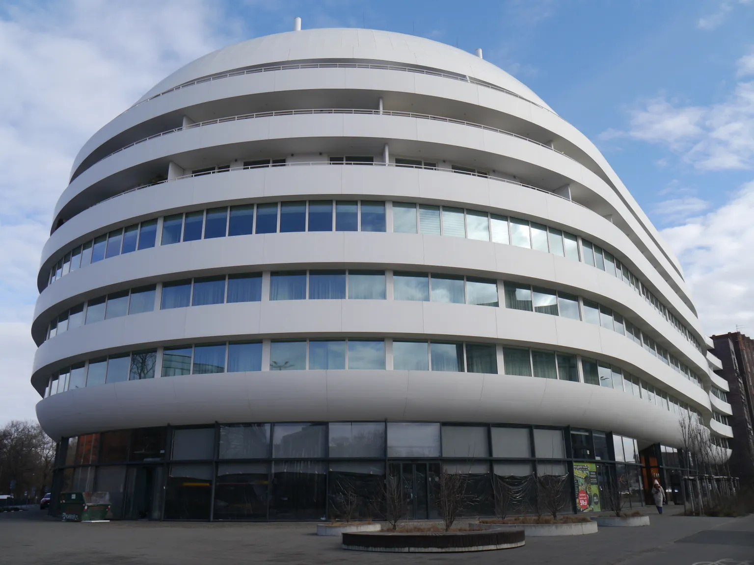 Modern, round, multi-story building - Ovo in Wrocław Poland, houses Pratia Wrocław,  with white, curved facade and numerous windows under a blue sky with clouds.