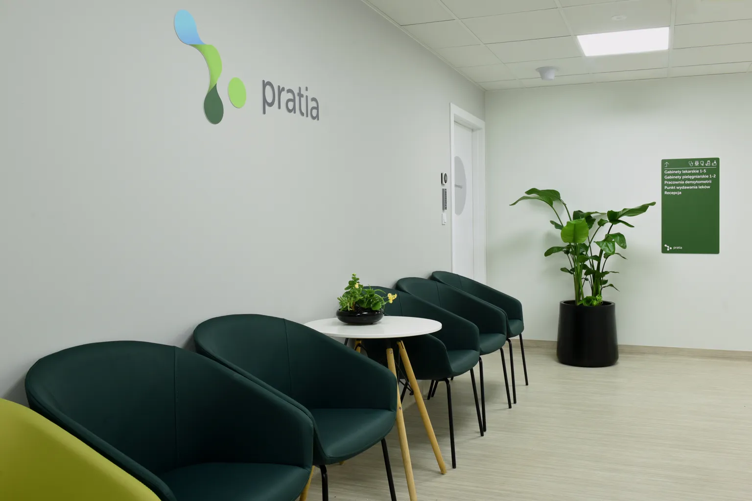Modern waiting room at a Pratia research site  with green chairs, a round table with a plant, wall sign and a Pratia logo, and a tall potted plant in the corner.